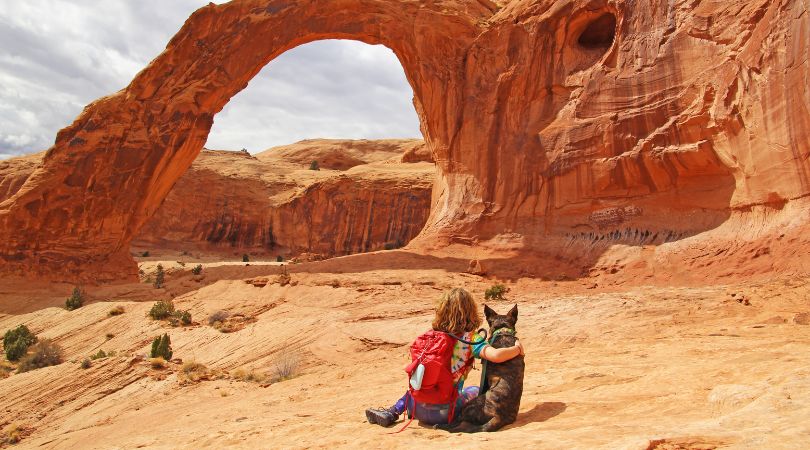 person with red backpack sitting with their arm around a dog looking at a red-rock arch