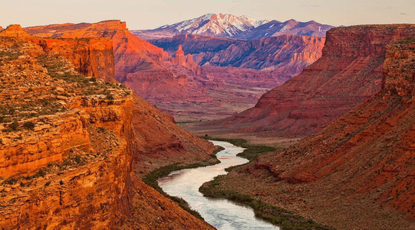 the colorado river flowing through red rock canyons with geological features like towers and buttes, and the la sal mountains in the background
